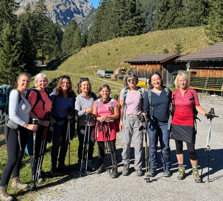 Gruppe von sieben Frauen mit Wanderstöcken in den Bergen vor einer Waldlandschaft.