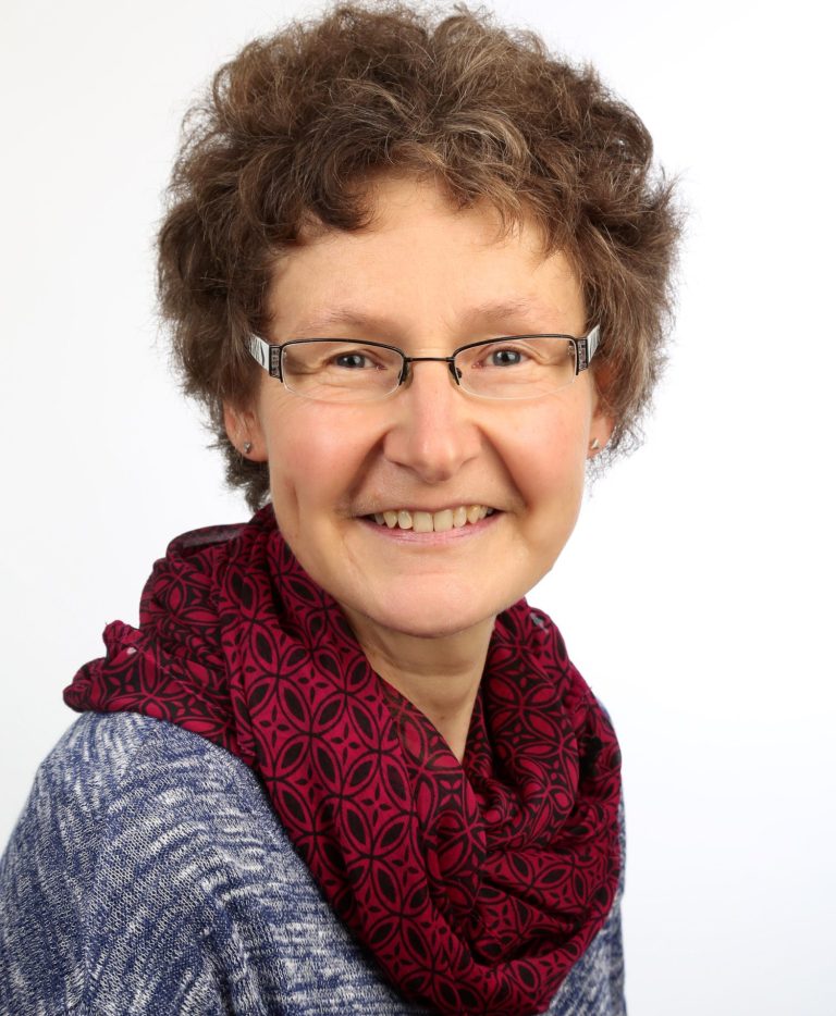 Frau mit lockigem Haar, Brille und rotem Schal, lächelnd vor hellem Hintergrund.